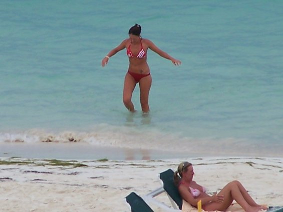 Woman in red and white bikini walks in shallow ocean water; another woman reclines on a beach chair