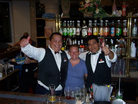 Two bartenders give thumbs up behind a bar stocked with bottles, with a customer between them