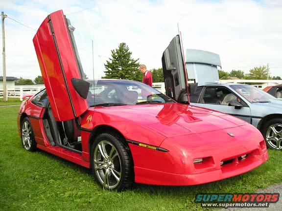 Red 1984 Pontiac Fiero with butterfly doors open, parked on grass next to a light blue car