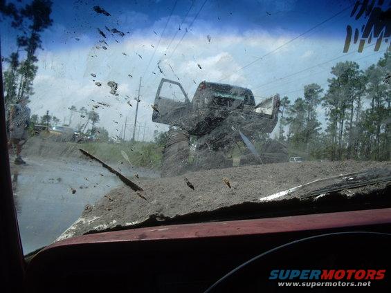 Mud-splattered monster truck with open doors stuck in mud, seen through a dirty windshield