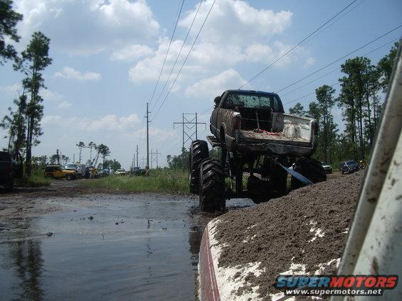 Muddy pickup truck with large tires in a mud pit, other vehicles and power lines in background