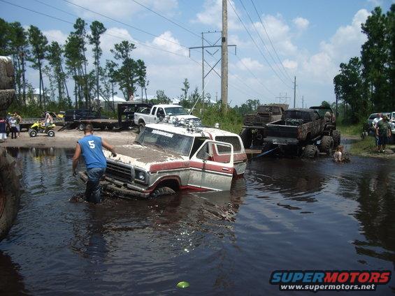 Mud-covered 1978 Ford Bronco partially submerged in water, with a man nearby