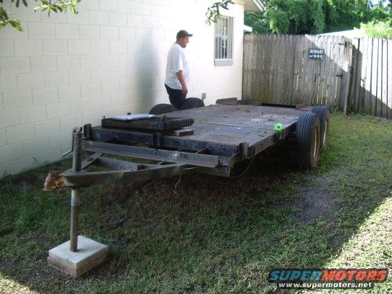 Flatbed trailer with two axles, a man standing nearby, and a wooden fence in the background