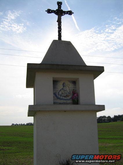 Roadside shrine with crucifix and religious imagery, set against a cloudy sky and green fields