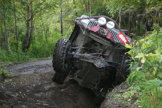 Off-road vehicle tilted on its side on a muddy trail with trees in the background. It has large tires and auxiliary lights on top
