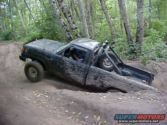 Muddy pickup truck stuck in a ditch on a wooded trail. It has a roll cage and a spare tire in the bed