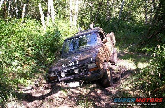 Pickup truck stuck in mud on a wooded trail. It has 'MUD ADDICT' and 'POORBOY OFFROAD' stickers