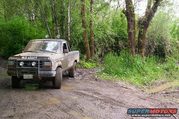 1992 Ford Ranger pickup truck covered in mud, with "MUD ADDICT" on the hood, driving on a muddy trail