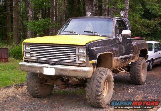 Lifted black pickup truck with yellow hood, large off-road tires, and a white car parked behind it