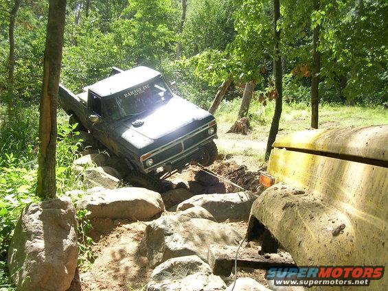 Dark pickup truck on rocky terrain, front grille visible, "DAMAGE INC" sticker on windshield