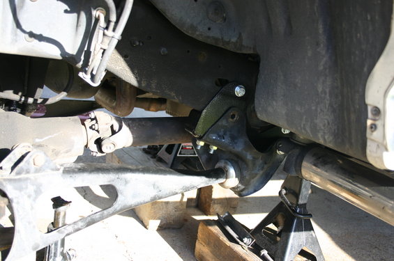 close-up of vehicle suspension parts on jack stands, showing a bracket bolted to the frame and a driveshaft