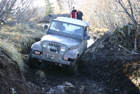 Jeep CJ-7 going down a muddy ditch with two people standing on a bank behind it