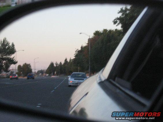View from a car's side mirror shows traffic on a road at dusk with trees lining the side