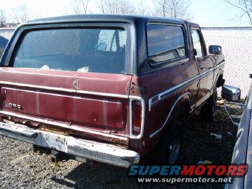 Maroon Ford Bronco with black top, seen from the back corner. Looks like it's in a junkyard