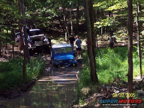 Blue Jeep Wrangler driving through a muddy stream in a forest, with another vehicle behind it