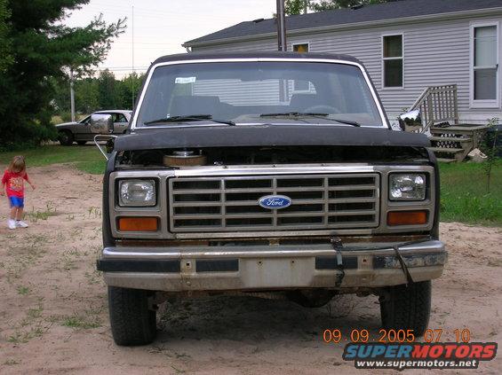 Front view of a black Ford Bronco with hood open. A child stands to the left. Another car is parked in the background