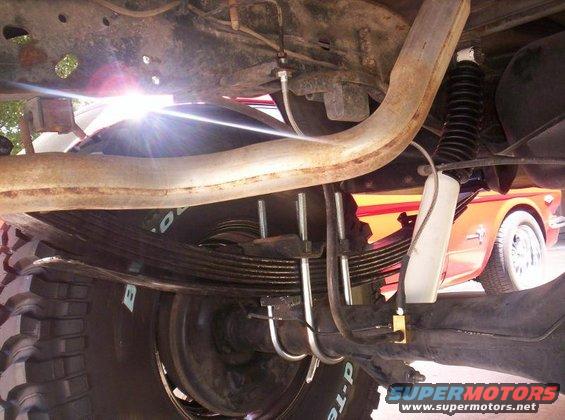 Underside view of a truck's rear leaf springs, U-bolts, shock absorber, and exhaust pipe. Another vehicle is visible in the background