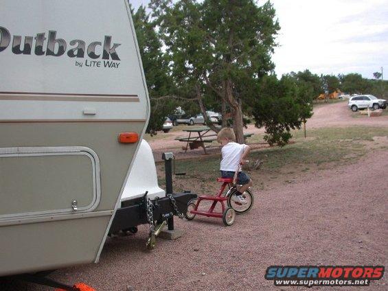 Child on red tricycle near trailer hitch, white SUV in background