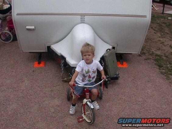 kid on a red tricycle, facing forward, in front of a white trailer hitch and landing gear
