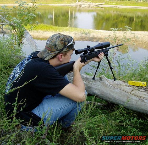 Person in camouflage hat and black t-shirt crouches by a log, aiming a rifle with a scope and bipod towards a body of water
