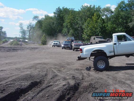 White pickup truck with large tires, side view, in a dusty outdoor area with other vehicles