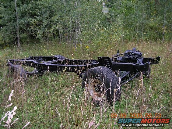 Abandoned vehicle frame with wheels and some drivetrain components, sitting in tall grass and weeds with trees in the background