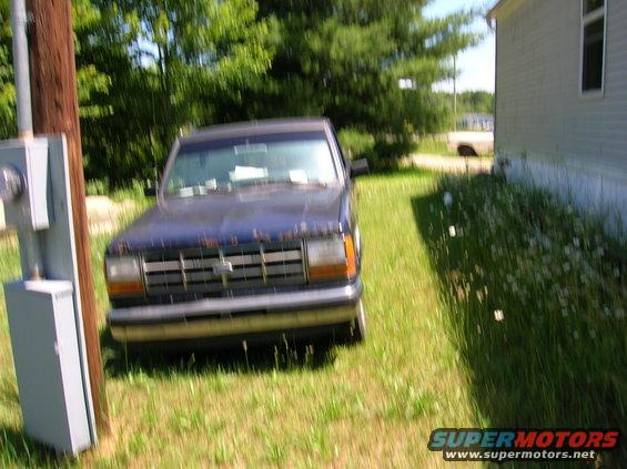 Blue pickup truck parked on grass next to a house. It has a horizontal grille and chrome bumper