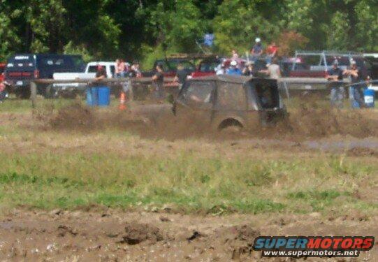 Muddy off-road vehicle driving through a mud pit with spectators in the background