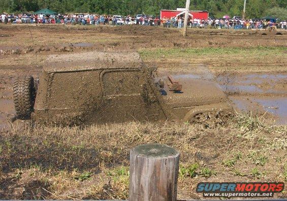 Mud-covered off-road vehicle stuck in a mud pit, side view. A spare tire is visible on the left