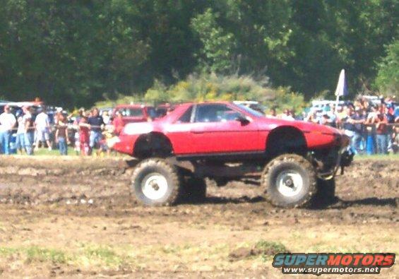 Red Pontiac Fiero modified with monster truck tires and suspension, driving in mud with a crowd watching