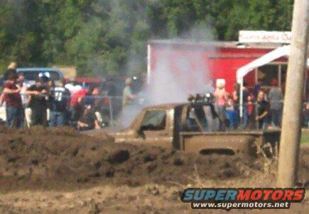 Mud-covered pickup truck stuck in mud, with people watching and smoke rising