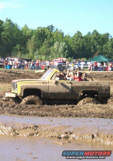 Mud-covered pickup truck with people in the cab stuck in a mud pit. Appears to be a 1973-1980 Chevrolet/GMC C/K series truck