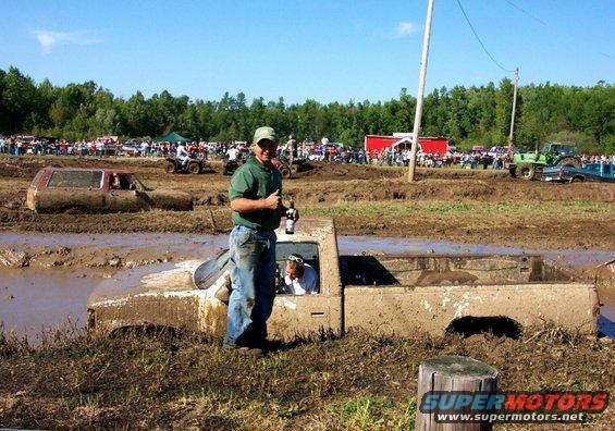 Mud-covered pickup truck stuck in a mud bog with a man standing next to it holding a beer