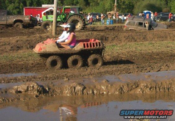 Six-wheeled amphibious vehicle in mud with two people riding