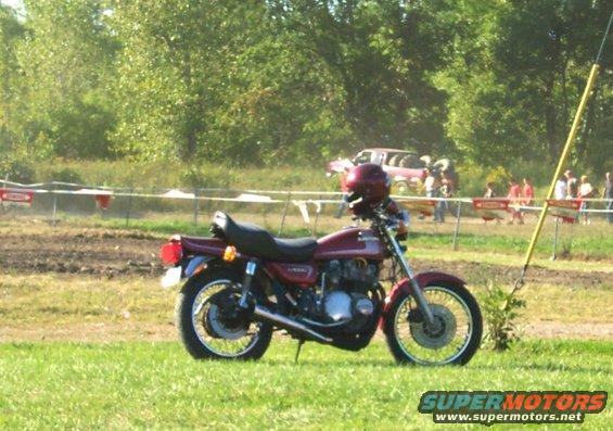 Maroon motorcycle parked on grass, helmet on handlebars. Dirt track and trees in background