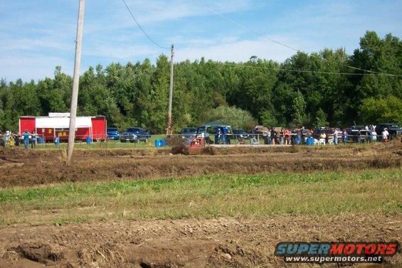 Mud bog scene with multiple trucks and spectators gathered near a red and white building