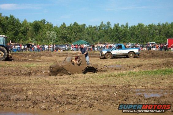 Trucks stuck in mud pit with spectators watching. One truck is partially submerged