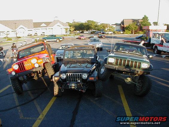 Three modified Jeeps parked at an angle in a parking lot, one orange, one black, and one green