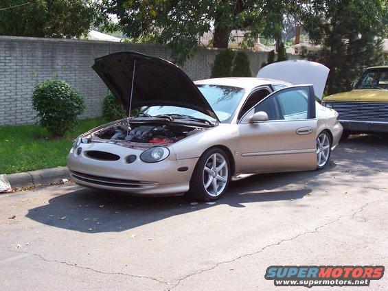 Tan sedan, hood and driver door open, parked next to a yellow truck. Looks like a 1996 model