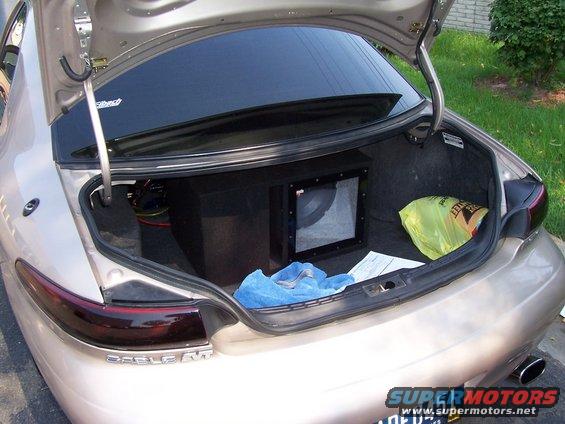 Rear view of a tan sedan trunk open, showing a subwoofer box and a yellow plastic bag