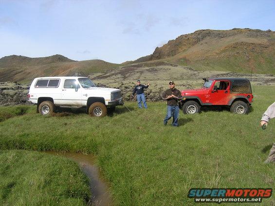 White Chevrolet K5 Blazer and red Jeep Wrangler parked in a field with people nearby