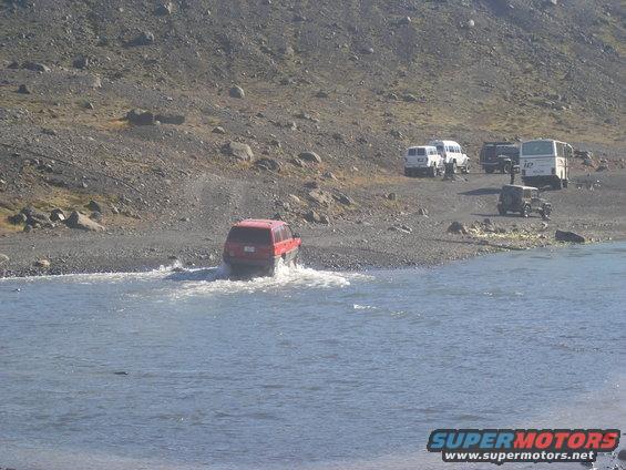 Red SUV driving through shallow water with a rocky hillside and other vehicles in the background