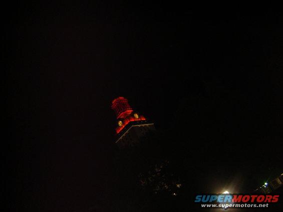 Dark scene with a tall building illuminated by red lights at night. A watermark reads SUPERMOTORS.NET