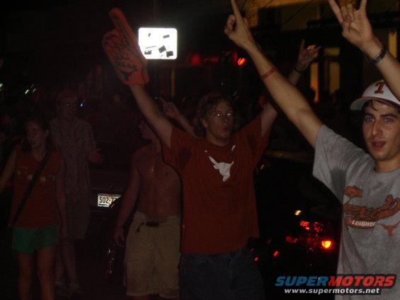 People with arms raised in celebration at night, one holding a large foam finger. A license plate is partially visible