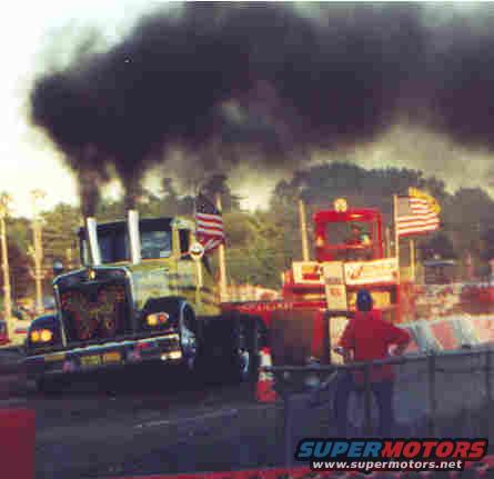Two trucks emitting black smoke during a pull competition, one green and one red
