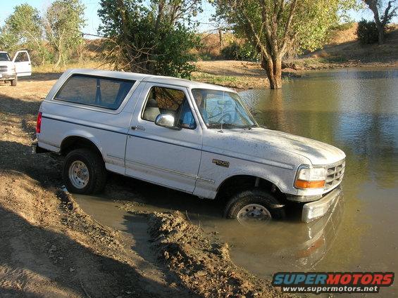 White Ford Bronco partially submerged in muddy water, front right wheel deep in mud. Another white SUV is parked nearby