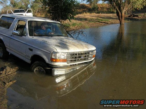 White 1994 Ford Bronco stuck in muddy water, front tires submerged. Another white vehicle visible behind it