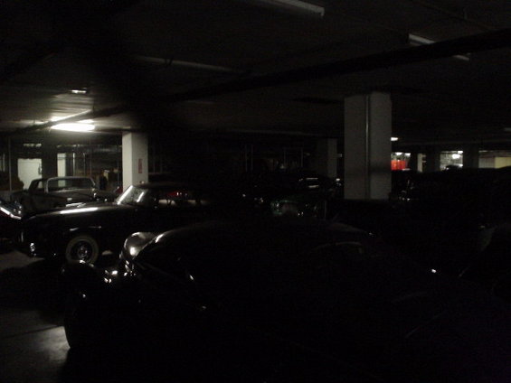 Several vintage cars parked in a dark garage, some with headlights on