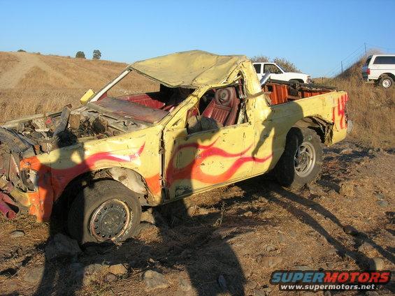 Wrecked yellow pickup truck with red and orange flame decals, flat front tire, and damaged roof. Two other white vehicles are visible in the background on a grassy hill