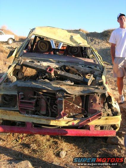 wrecked yellow vehicle with red bumper, person standing next to it. White SUV in background